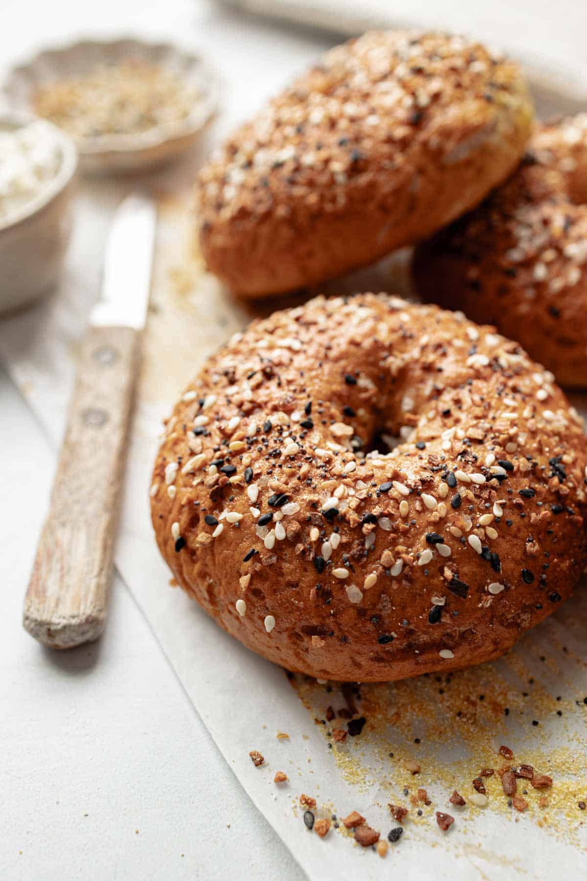 A close-up shot of a gluten-free bagel with a crispy, golden crust covered in everything bagel seasoning, with a knife and small bowl of seasoning nearby.