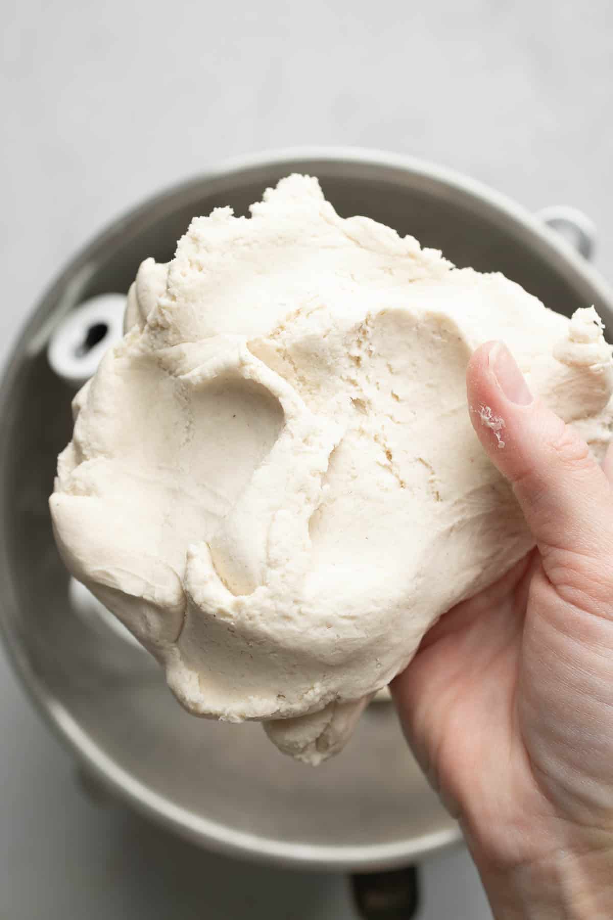 A close-up of a hand holding a smooth, elastic ball of gluten-free bagel dough above a mixing bowl, showing its texture and consistency.
