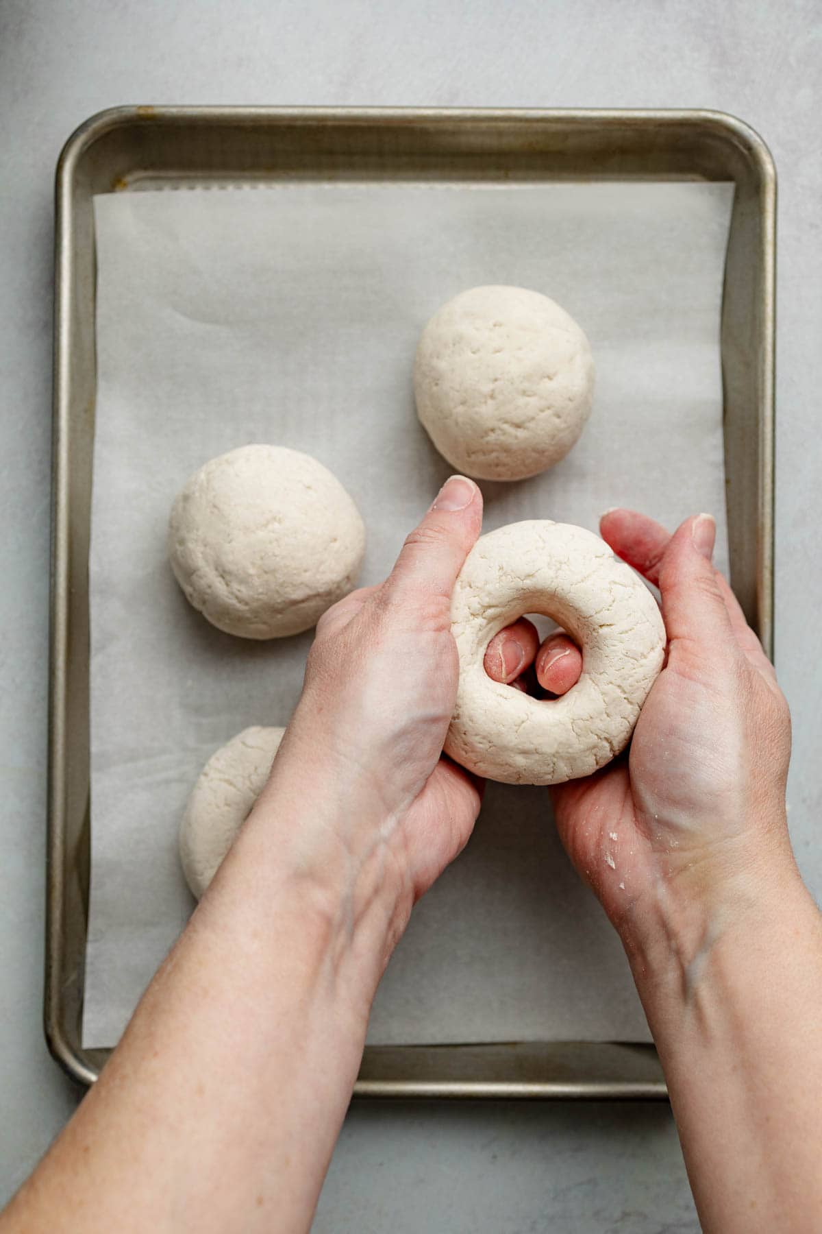 Hands shaping a gluten-free bagel by pressing fingers through the center of a dough ball. Other shaped dough balls rest on a parchment-lined baking sheet.