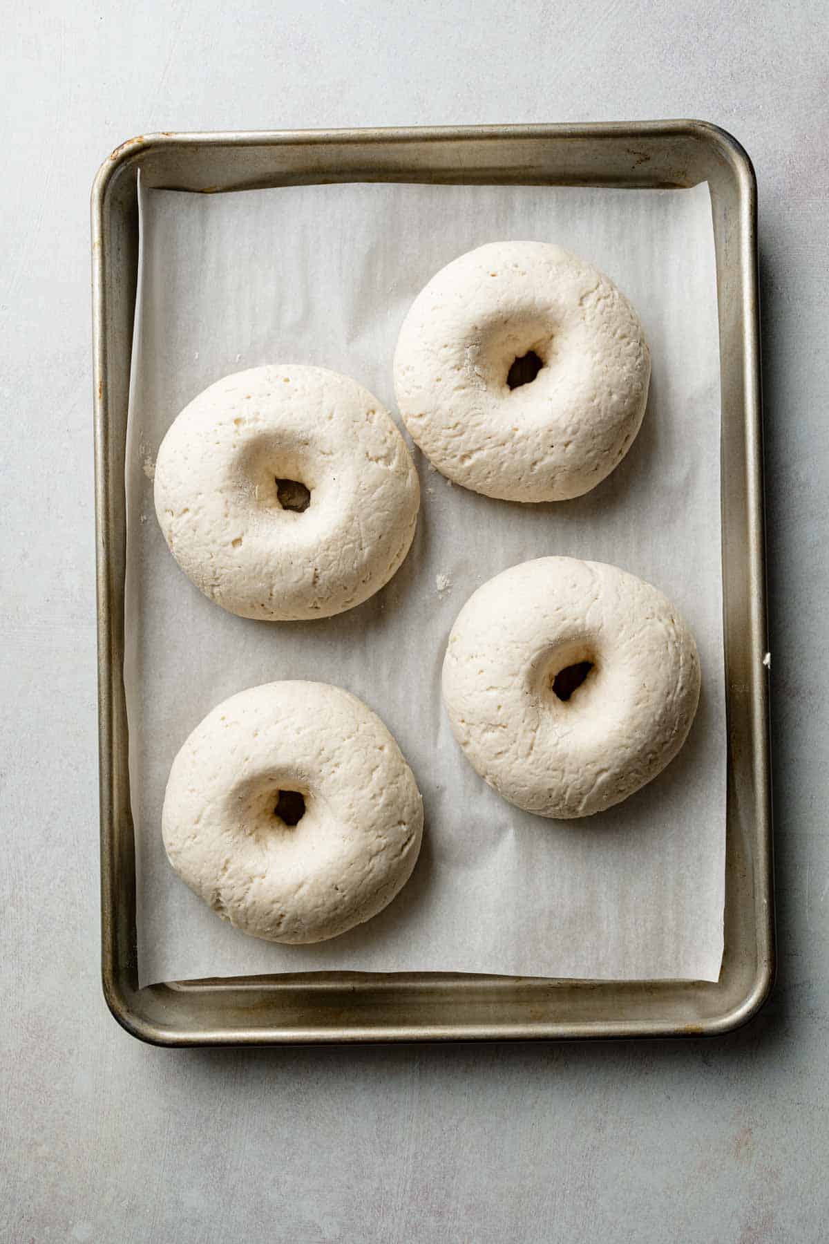 Four gluten-free bagels resting on a parchment-lined baking sheet after proofing. The dough has expanded slightly, looking soft and airy, ready for boiling.