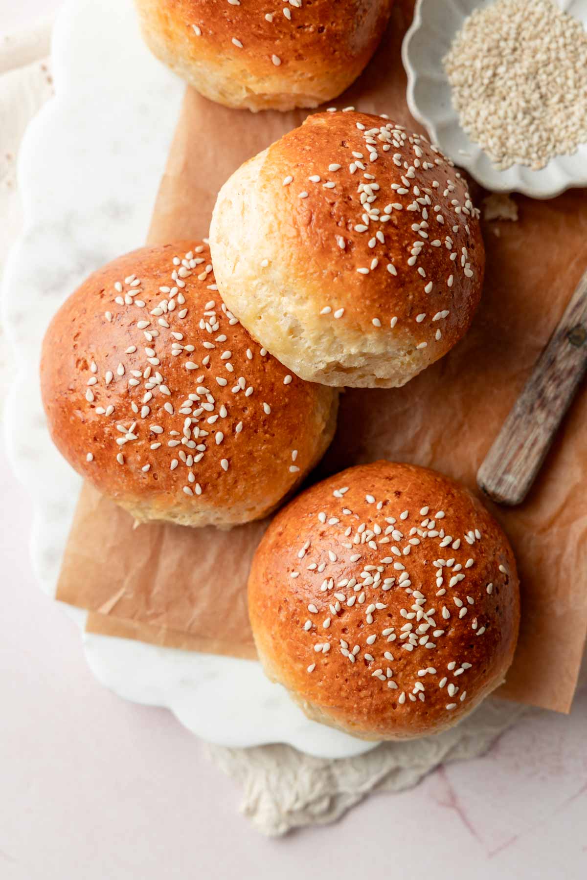Overhead view of golden-brown gluten-free hamburger buns topped with sesame seeds, arranged on parchment paper with a small dish of sesame seeds nearby.