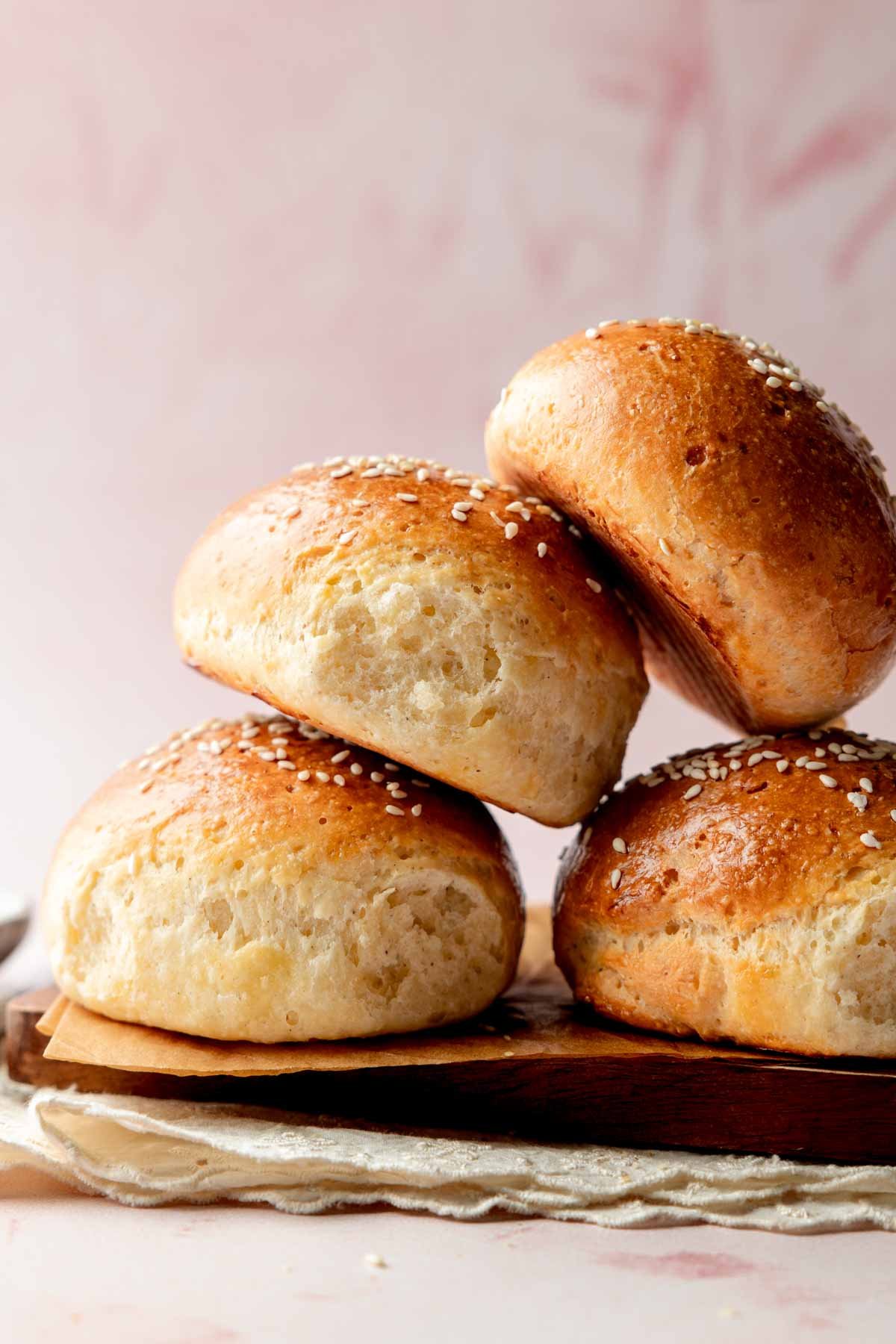 Side view of a stack of fluffy gluten-free hamburger buns with shiny, golden crusts and sesame seed topping, placed on a wooden board with a soft pink background.