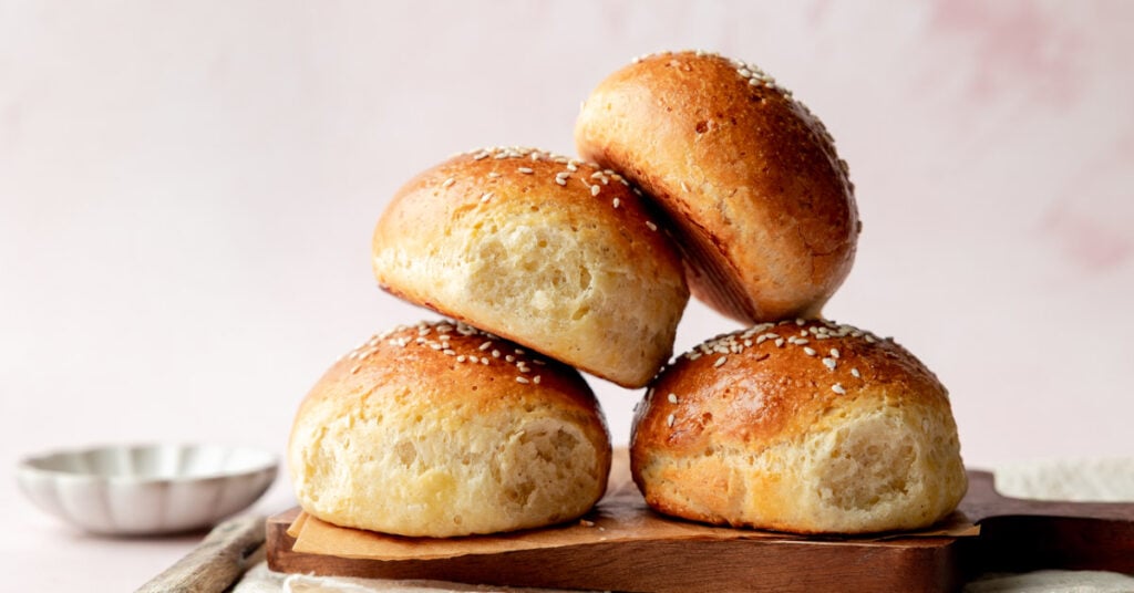 Side view of a stack of fluffy gluten-free hamburger buns with shiny, golden crusts and sesame seed topping, placed on a wooden board with a soft pink background.