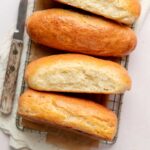 Overhead view of four gluten-free hot dog buns on a cooling rack with parchment paper, two of them cut to reveal a soft and airy crumb.
