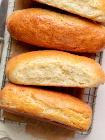 Overhead view of four gluten-free hot dog buns on a cooling rack with parchment paper, two of them cut to reveal a soft and airy crumb.
