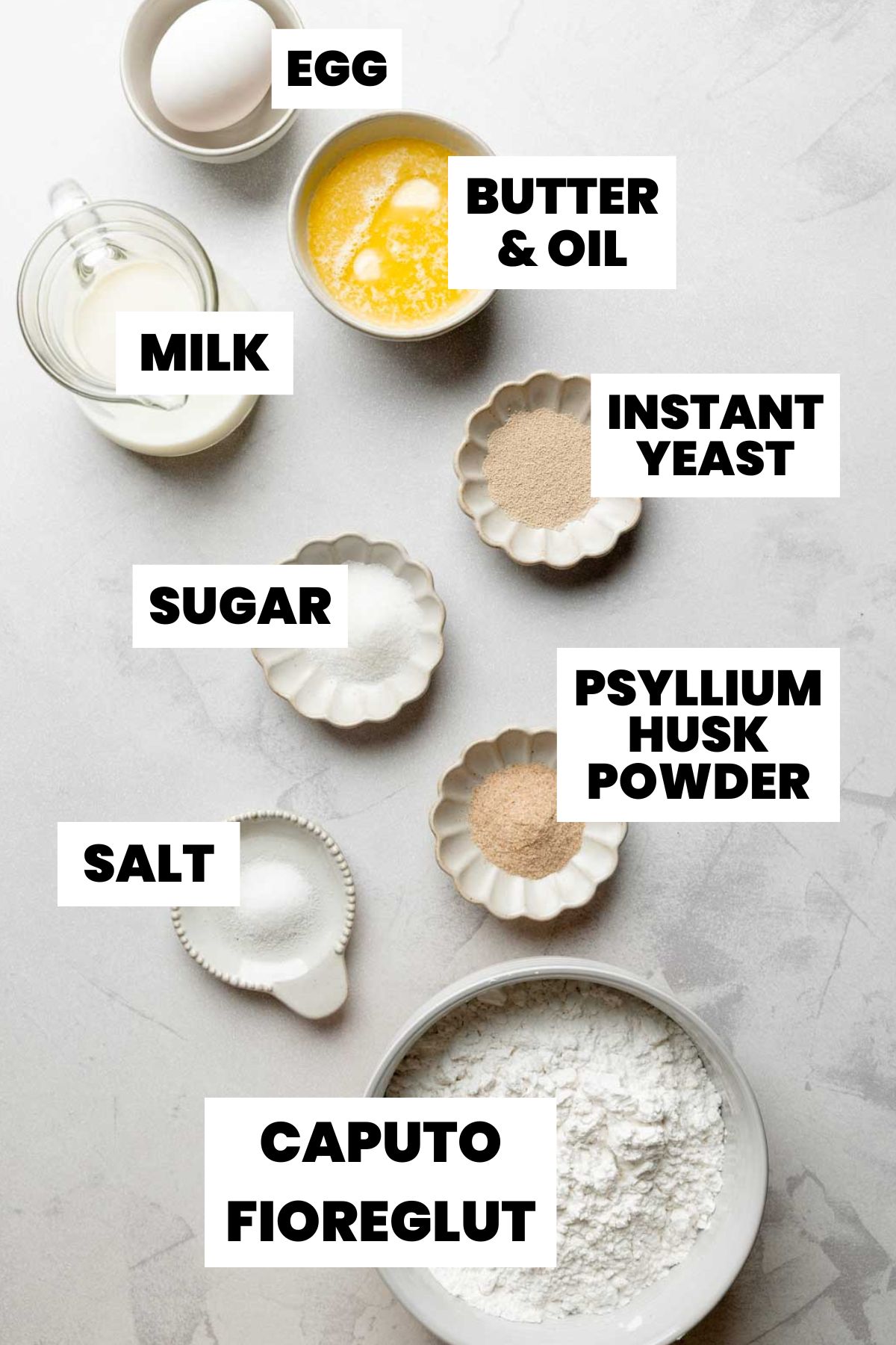 Overhead view of labeled ingredients for gluten-free hamburger and hot dog buns, including flour, yeast, milk, butter, egg, sugar, salt, and psyllium husk powder.