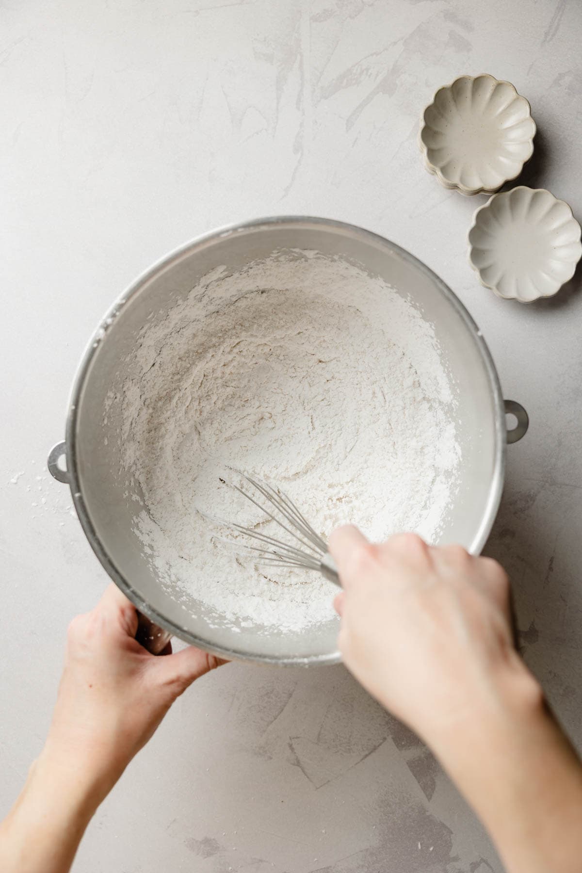 Hands whisking dry ingredients in a large metal mixing bowl on a light gray surface.
