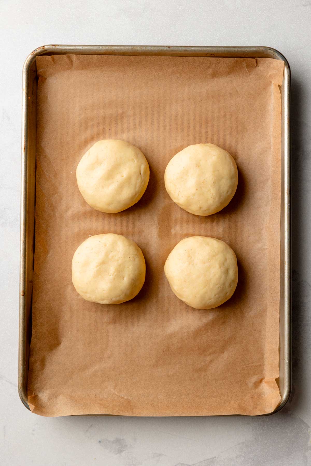 Four portions of gluten-free dough shaped into round buns, placed on a parchment-lined baking sheet before rising.