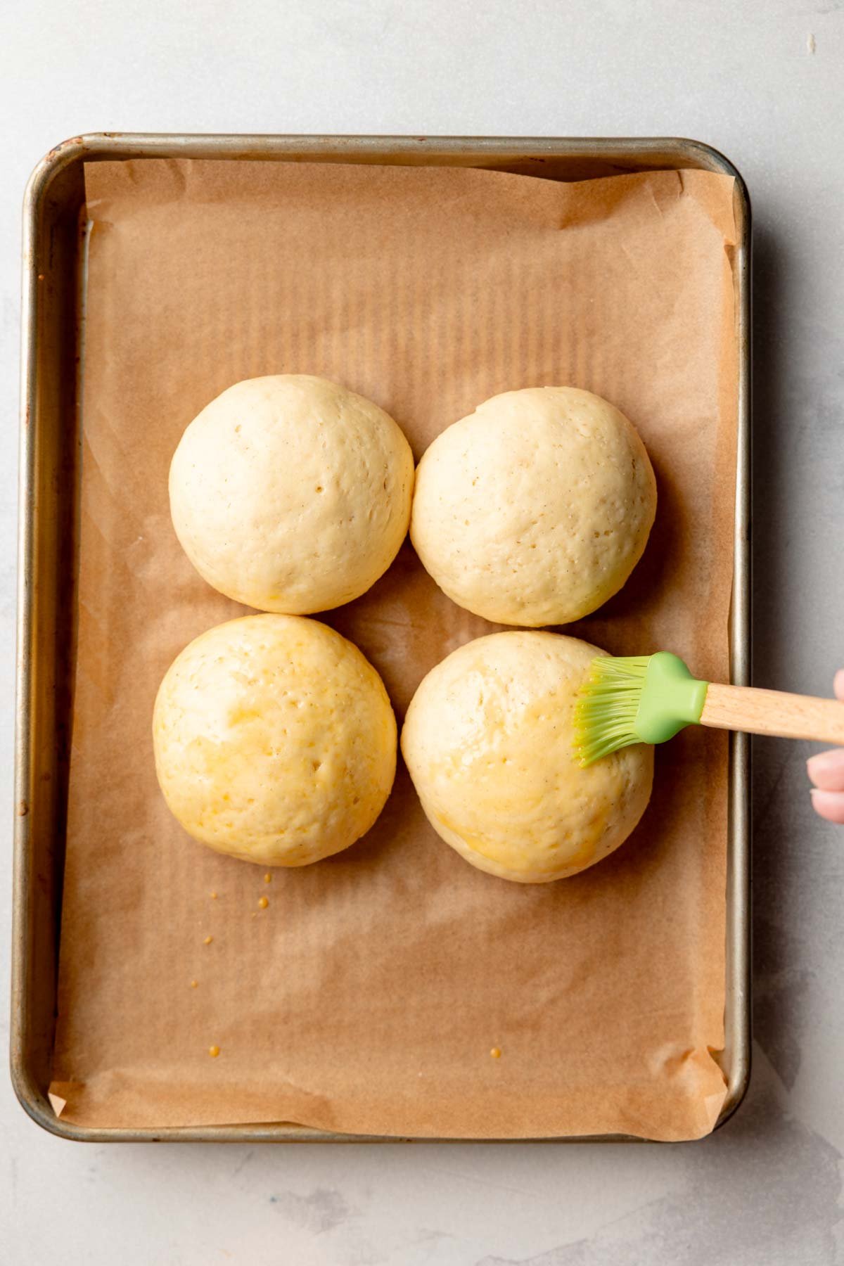 Gluten-free buns being brushed with egg wash using a green silicone pastry brush on a parchment-lined pan.
