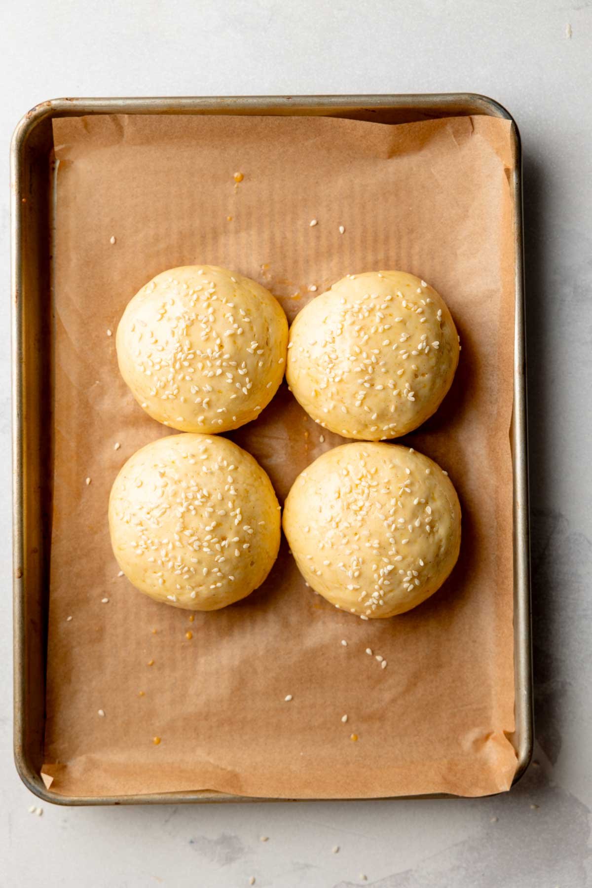Risen and egg-washed gluten-free buns sprinkled with sesame seeds on a parchment-lined baking sheet, ready to bake.