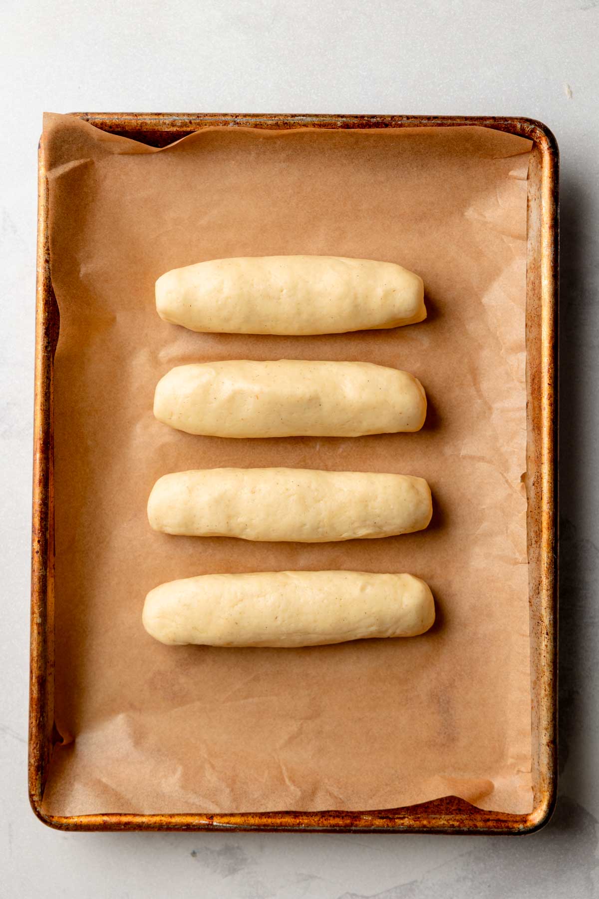 Four shaped gluten-free hot dog buns arranged on a parchment-lined baking sheet before rising.