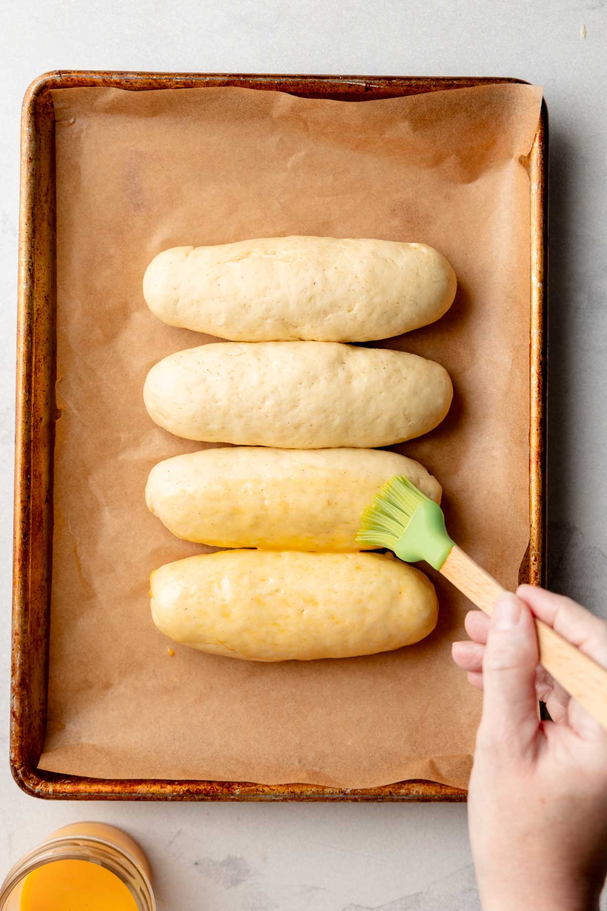Hand brushing egg wash onto risen gluten-free hot dog buns using a green silicone pastry brush.