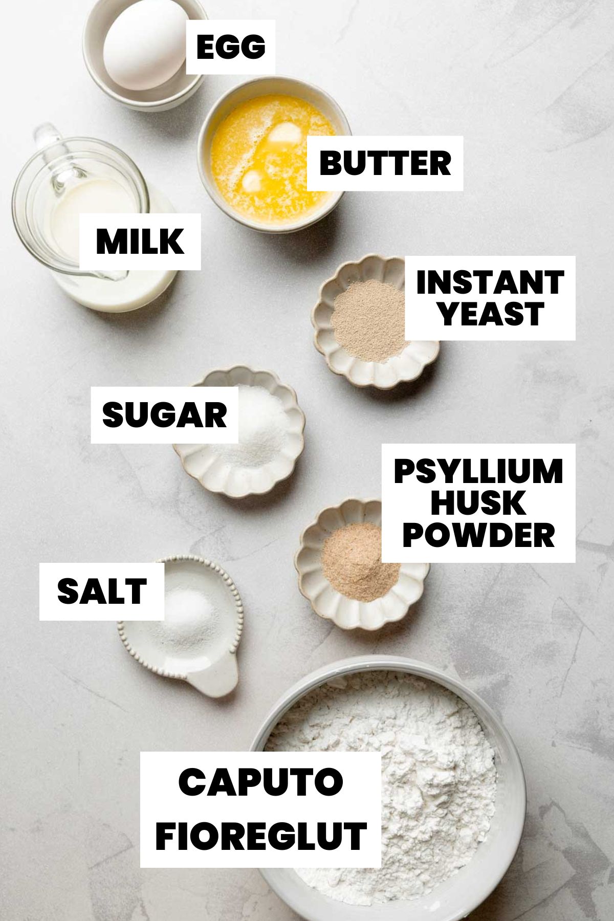 Overhead view of labeled ingredients for gluten-free hamburger and hot dog buns, including flour, yeast, milk, butter, egg, sugar, salt, and psyllium husk powder.