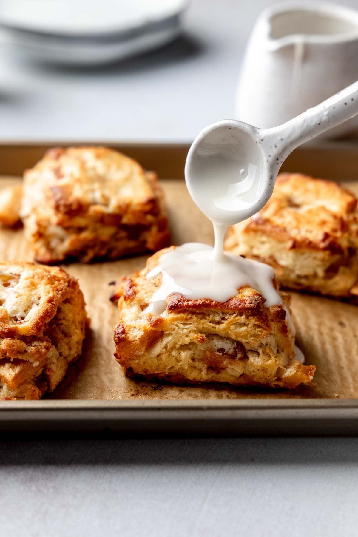 Close-up of glaze being spooned over a freshly baked apple biscuit on a parchment-lined baking tray.