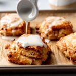 Spoon drizzling glaze over a warm gluten-free apple biscuit on a baking sheet with more biscuits in the background.