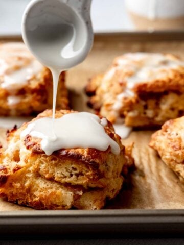 Spoon drizzling glaze over a warm gluten-free apple biscuit on a baking sheet with more biscuits in the background.