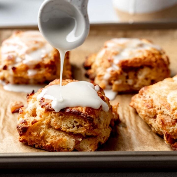Spoon drizzling glaze over a warm gluten-free apple biscuit on a baking sheet with more biscuits in the background.