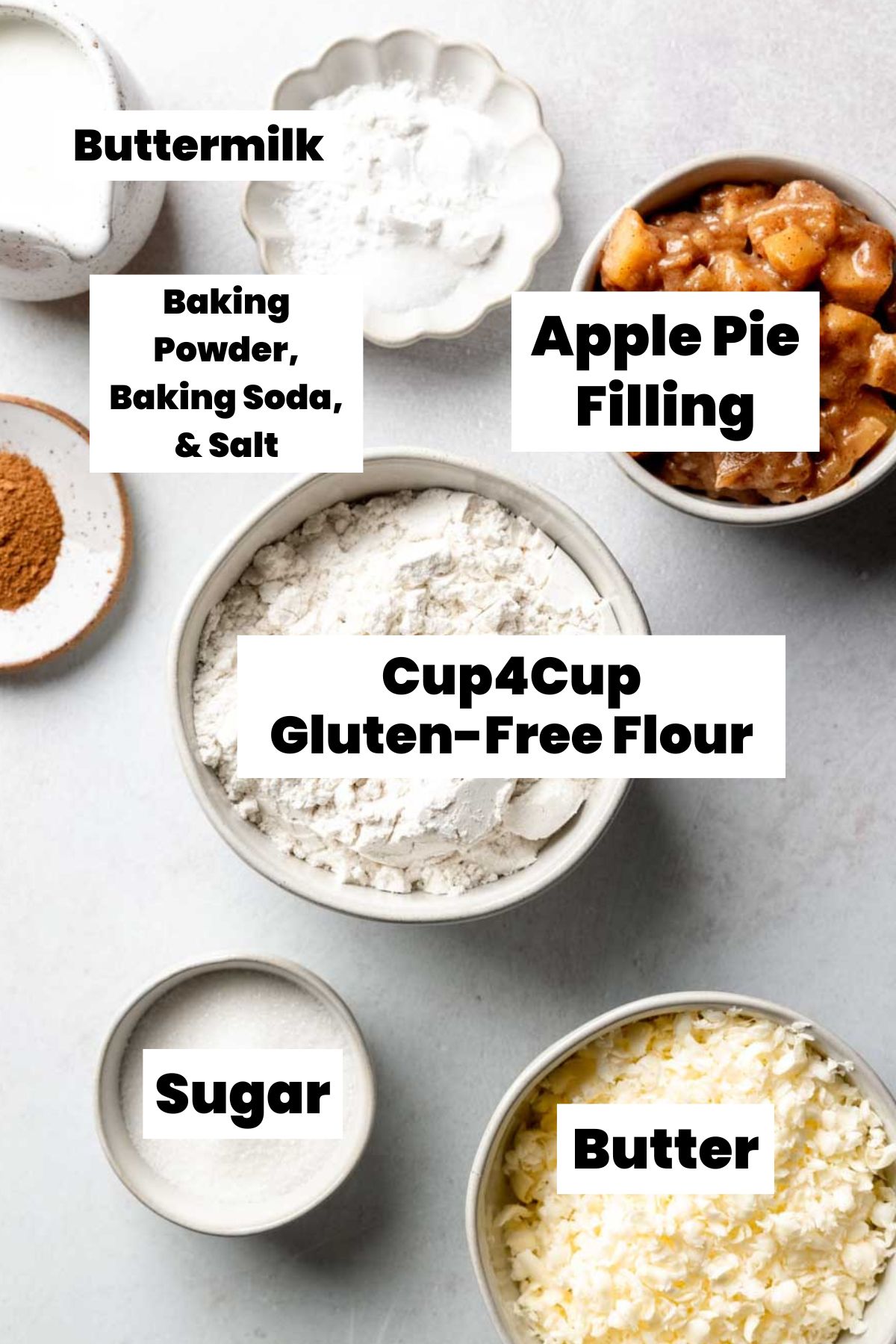 Flat lay of labeled gluten-free apple biscuit ingredients in bowls, including buttermilk, baking powder, baking soda, salt, apple pie filling, Cup4Cup gluten-free flour, sugar, and grated butter.