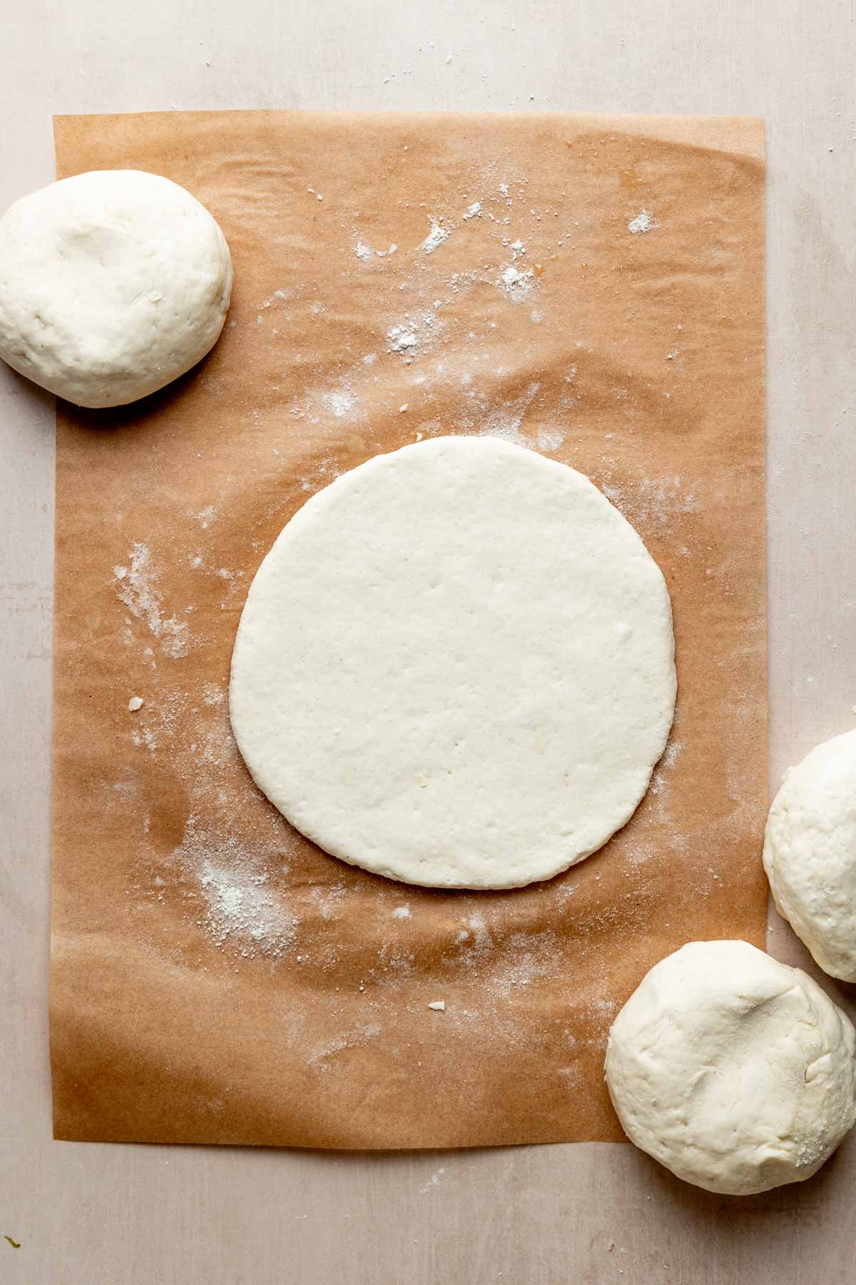 One ball of dough is rolled out into a disc on parchment paper, resting next to other balls of dough.