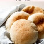 Puffed pita bread with golden top is in a cloth napkin lined bowl, stacked in front of the other pita breads.