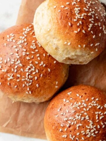 Overhead view of golden-brown gluten-free hamburger buns topped with sesame seeds, arranged on parchment paper with a small dish of sesame seeds nearby.