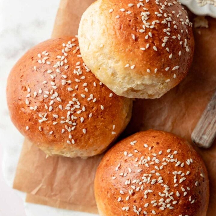 Overhead view of golden-brown gluten-free hamburger buns topped with sesame seeds, arranged on parchment paper with a small dish of sesame seeds nearby.