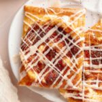 Close-up of a single pumpkin danish on a white speckled plate. The flaky crust and pumpkin filling are topped with a neat drizzle of icing.