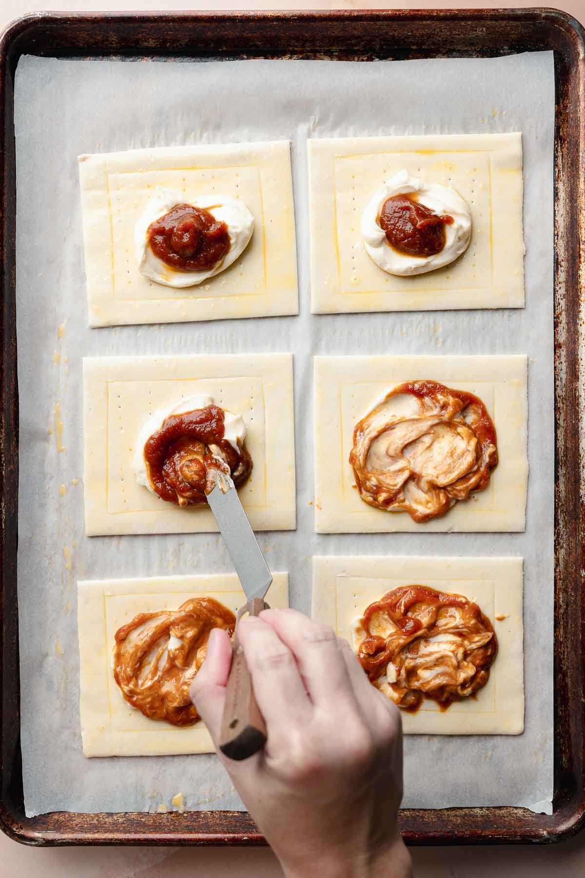 Unbaked pastries being assembled. Each puff pastry rectangle has a dollop of cream cheese topped with pumpkin mixture. A hand spreads the filling on one.