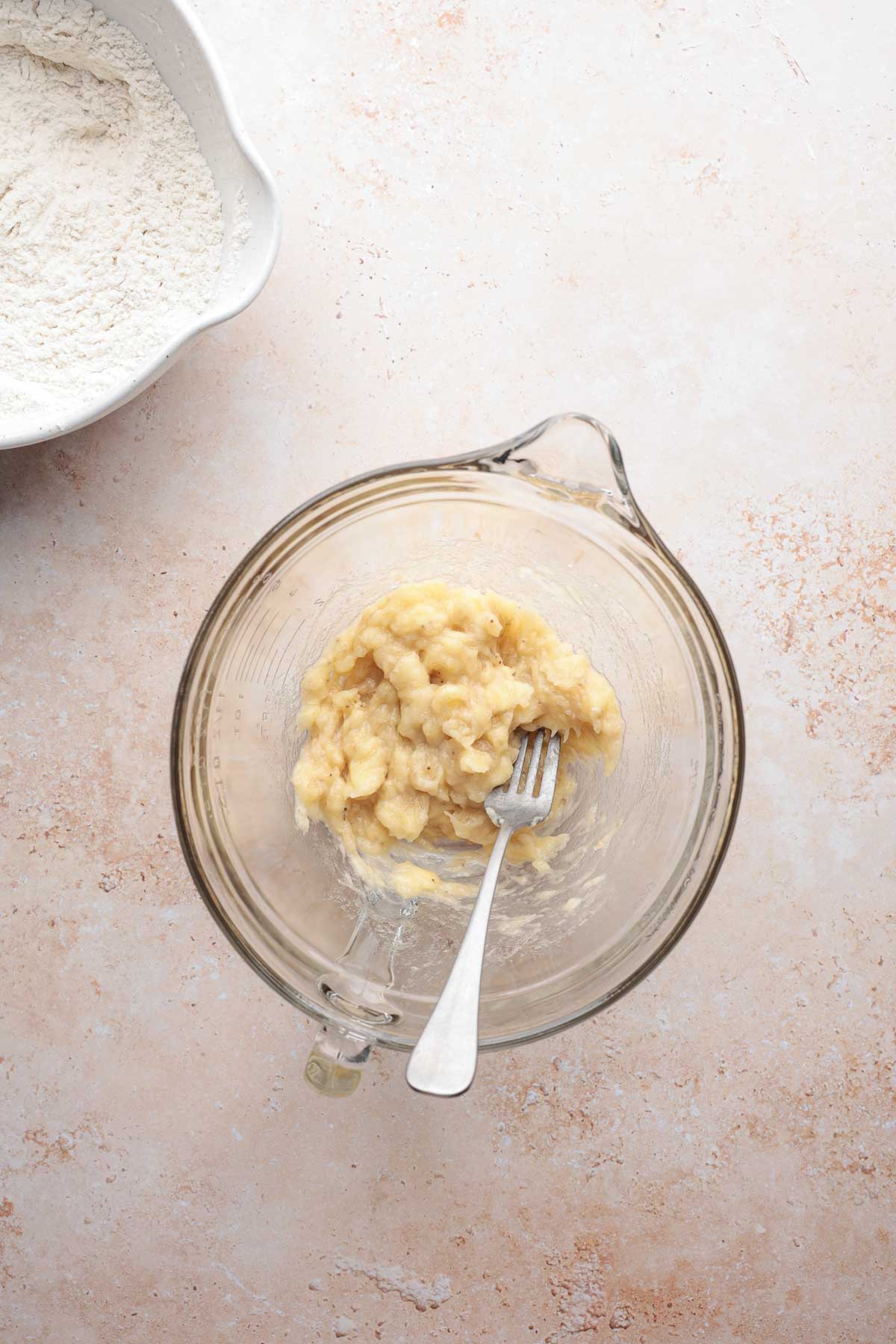 Mashed bananas in glass bowl beside flour.