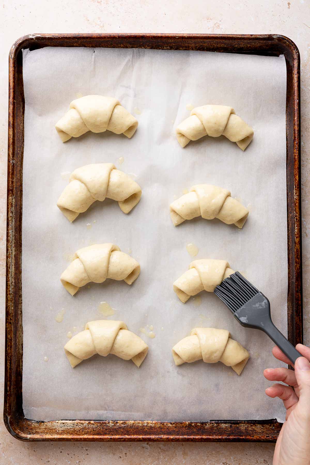 Baking sheet with unbaked gluten-free crescent rolls being brushed with butter.