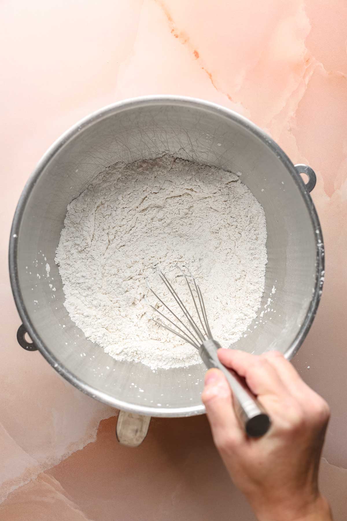 Dry ingredients in large stand mixer bowl, hand holding whisk in bowl.