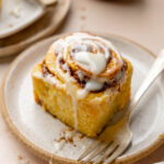 Gluten free pumpkin cinnamon roll on a plate with fork, icing drizzling down side.