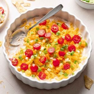 A white casserole dish layered with cream cheese taco dip base, cheddar cheese, orange bell pepper, and green onion; spoon resting in scooped out area.