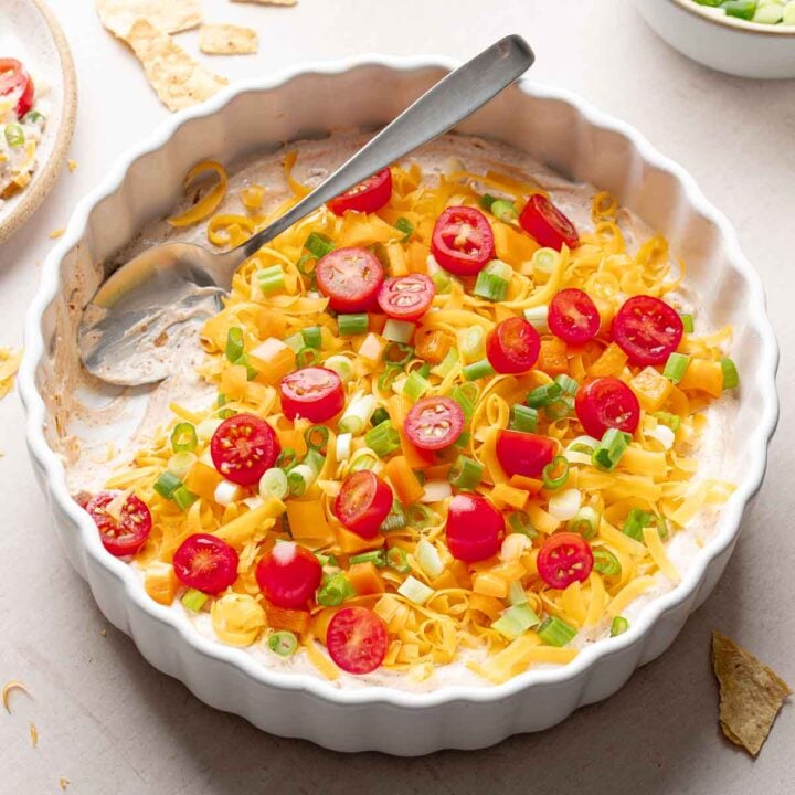 A white casserole dish layered with cream cheese taco dip base, cheddar cheese, orange bell pepper, and green onion; spoon resting in scooped out area.