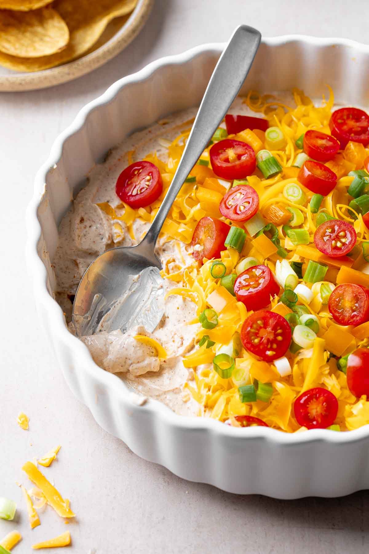 A white casserole dish layered with cream cheese taco dip base, cheddar cheese, orange bell pepper, and green onion; spoon resting in scooped out area.