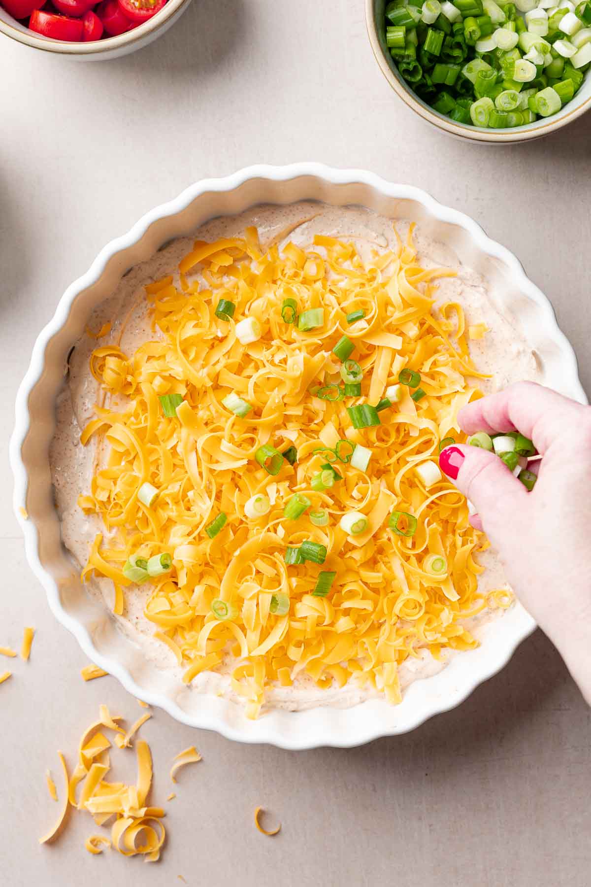 A white casserole dish layered with cream cheese taco dip base and cheddar cheese, hand sprinkling green onions over top.