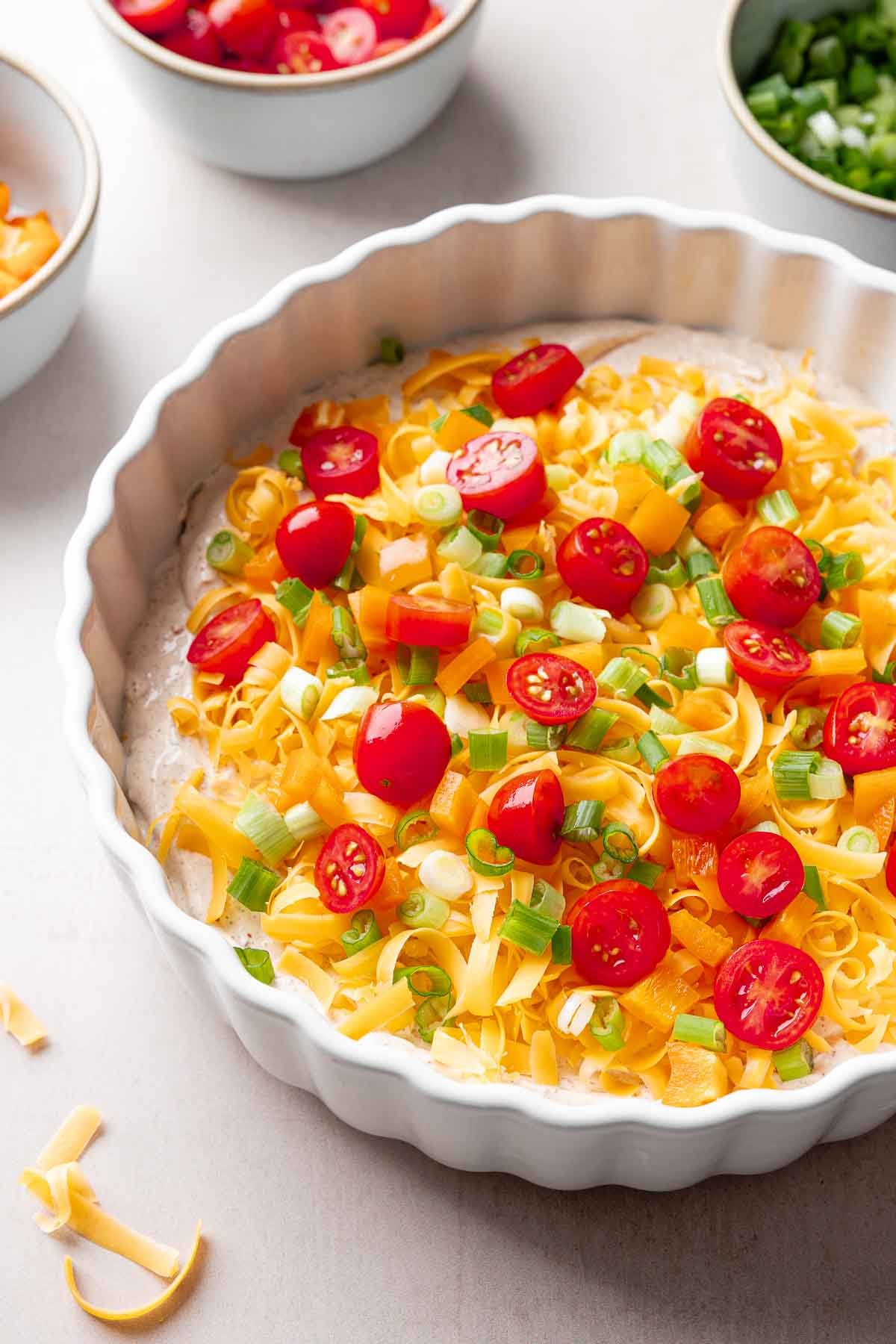 A white casserole dish layered with cream cheese taco dip base, cheddar cheese, orange bell pepper, and green onion.