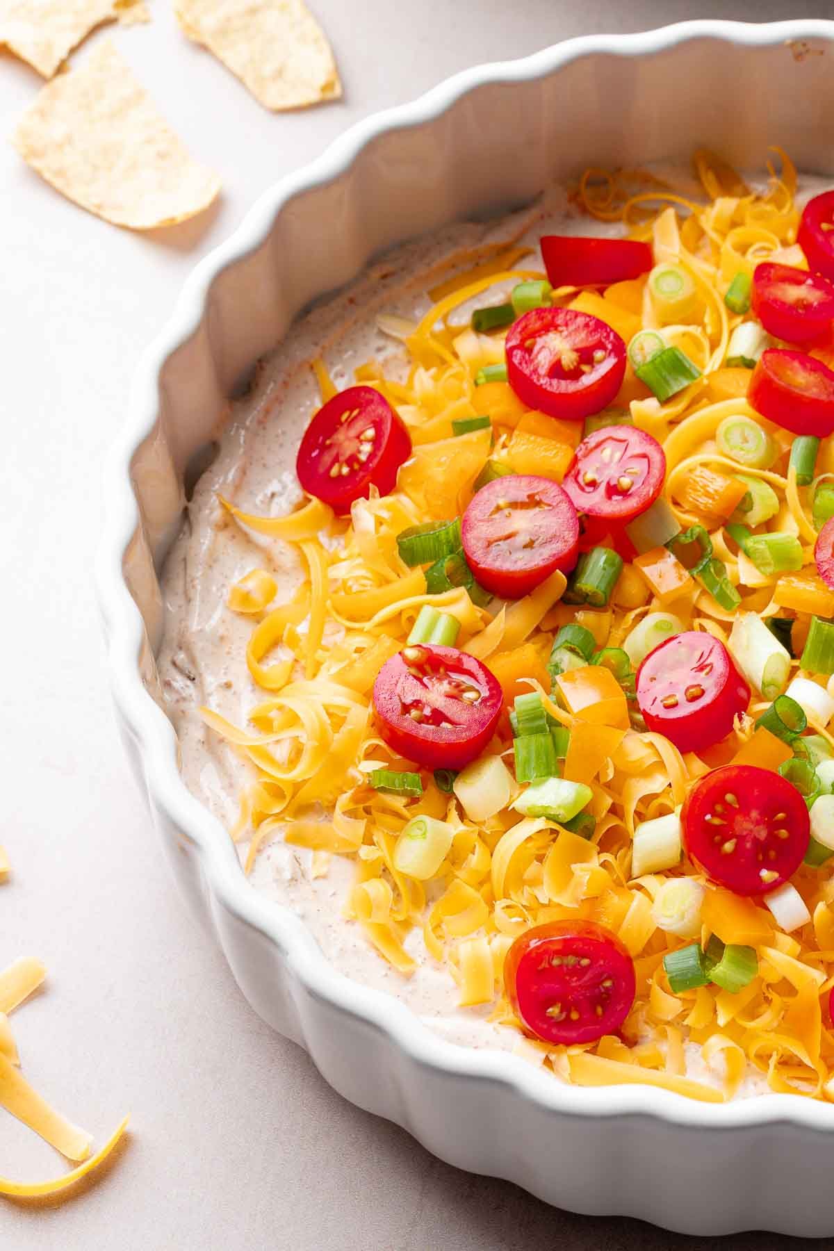 A white casserole dish layered with cream cheese taco dip base, cheddar cheese, orange bell pepper, and green onion.