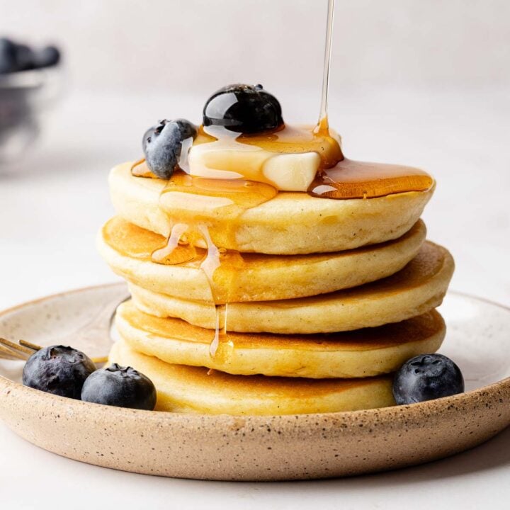 Syrup drizzling over a stack of fluffy gluten-free pancakes, garnished with a pat butter and blueberries.