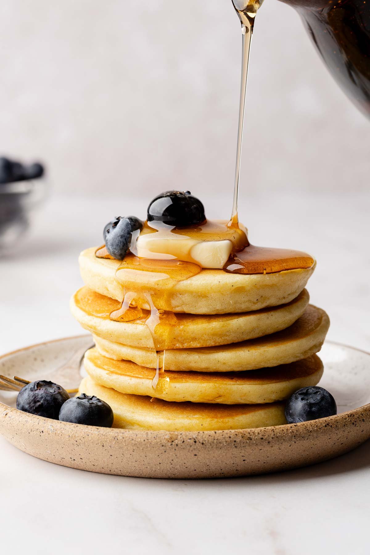 Syrup drizzling over a stack of fluffy gluten-free pancakes, garnished with a pat butter and blueberries.