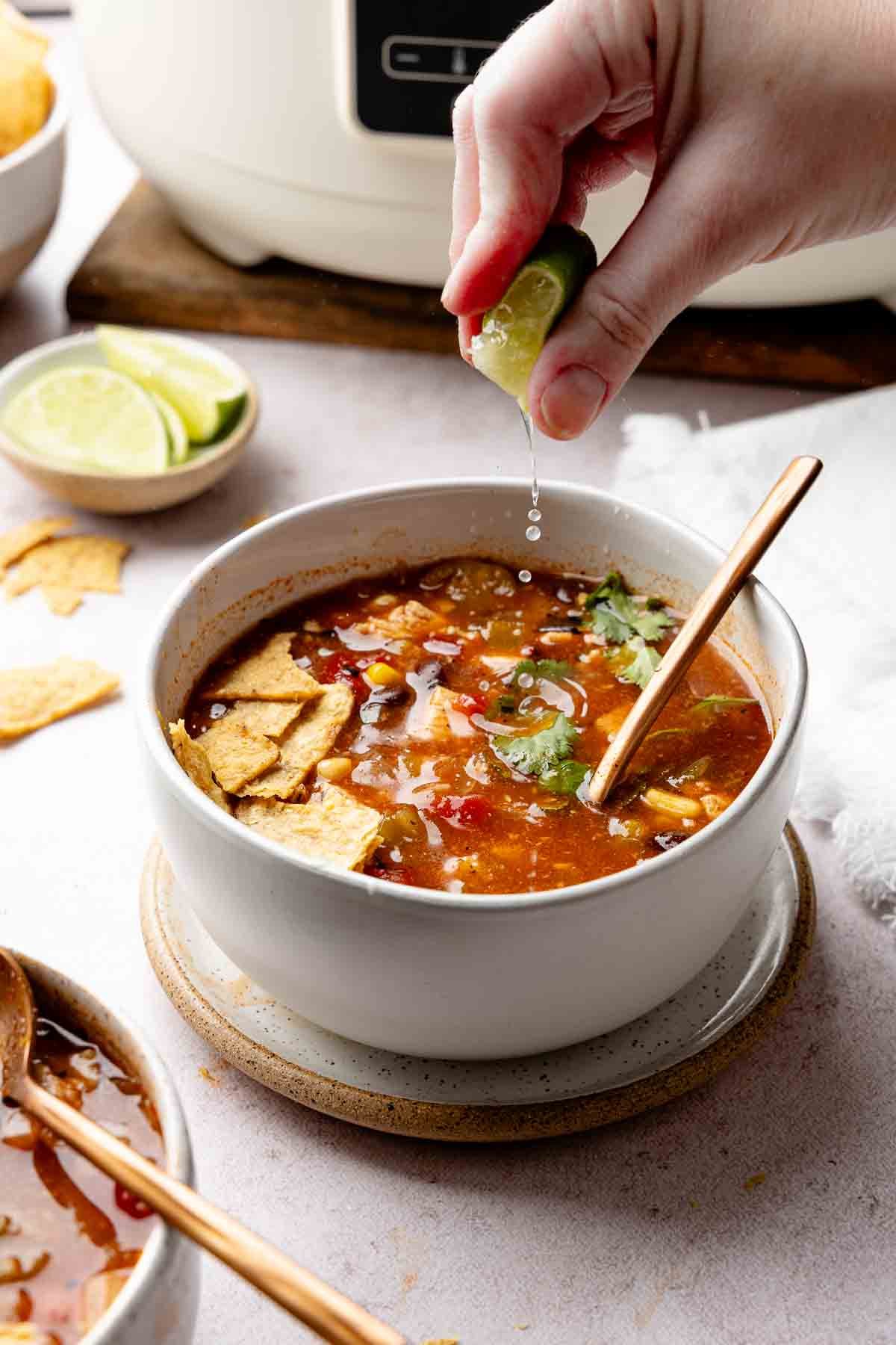 Hand squeezing lime juice into bowl of chicken tortilla soup garnished with tortilla chips.