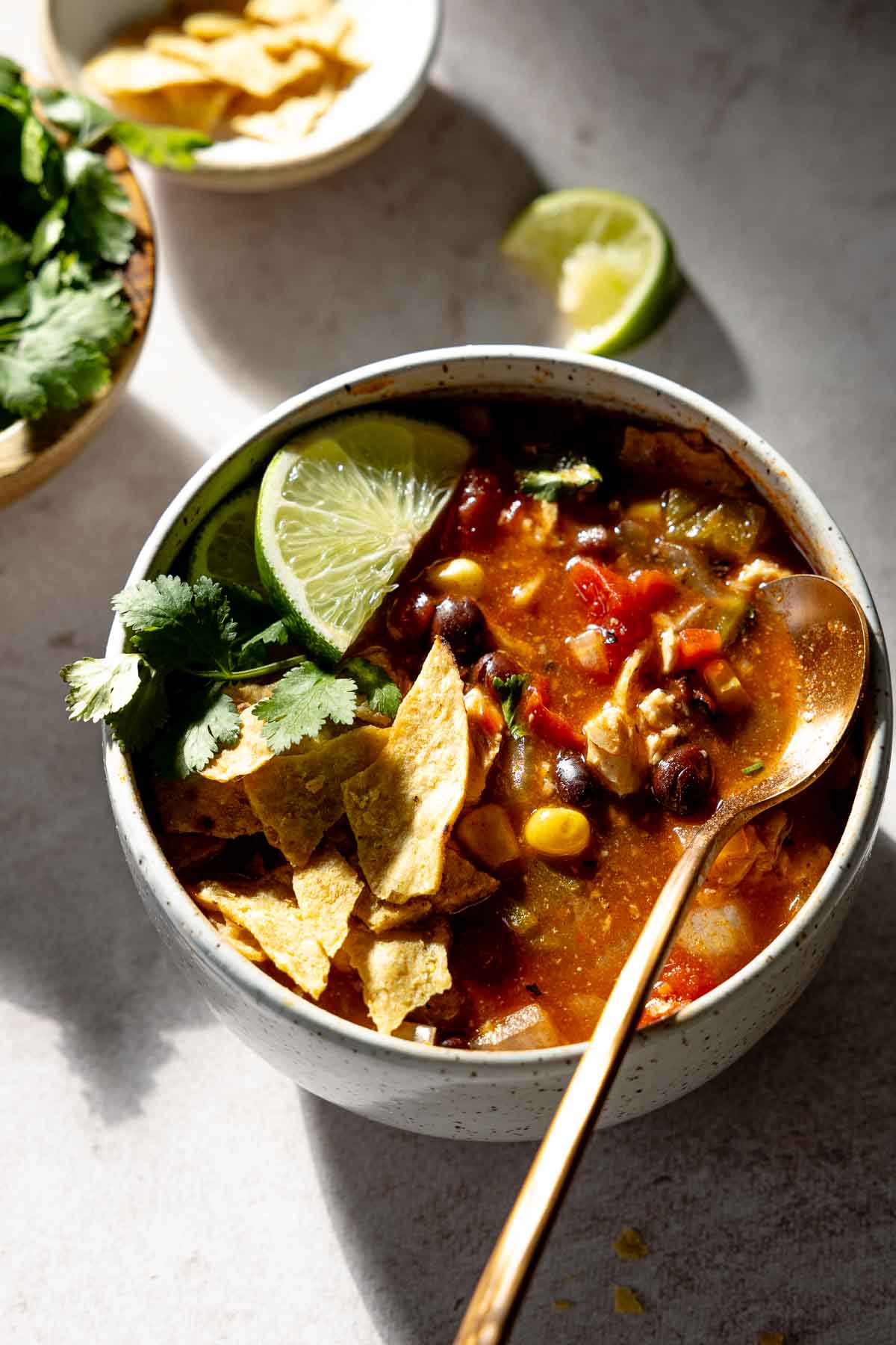 Chicken tortilla soup in bowl, with spoon resting to side, garnished with lime, cilantro, and tortilla chips.