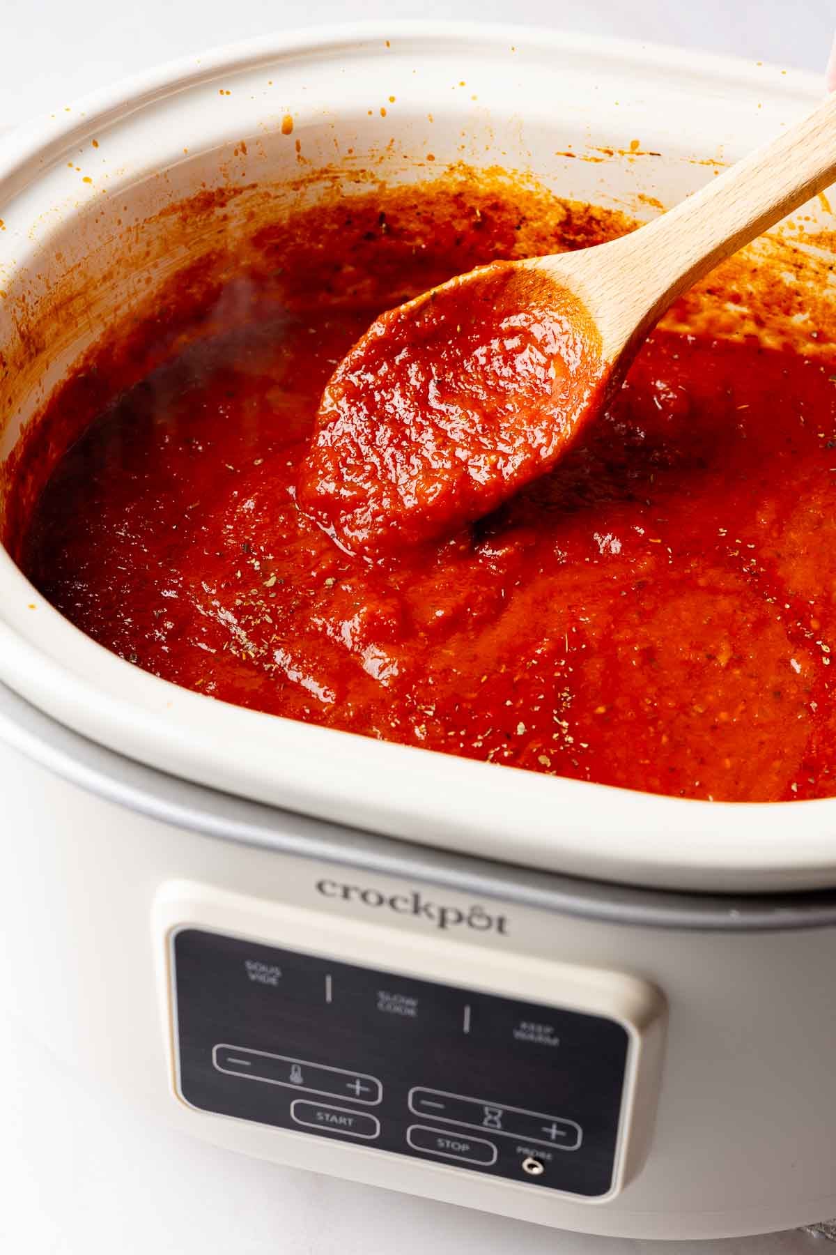 Overhead view of rich red spaghetti sauce in a white slow cooker with a wooden spoon.