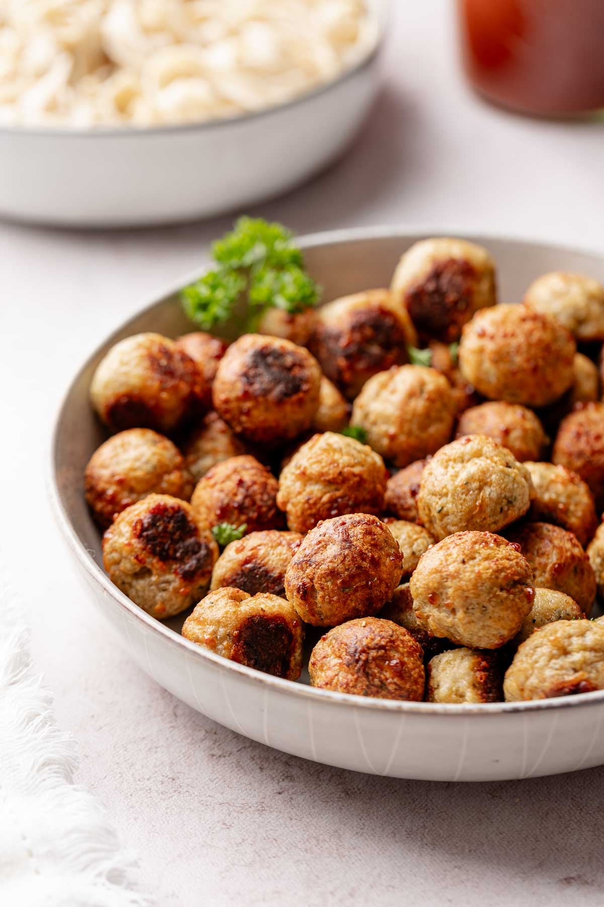 A large shallow white bowl filled with golden-brown baked turkey meatballs, garnished with a sprig of curly parsley. A blurred bowl of pasta and a jar of marinara sauce are visible in the background.