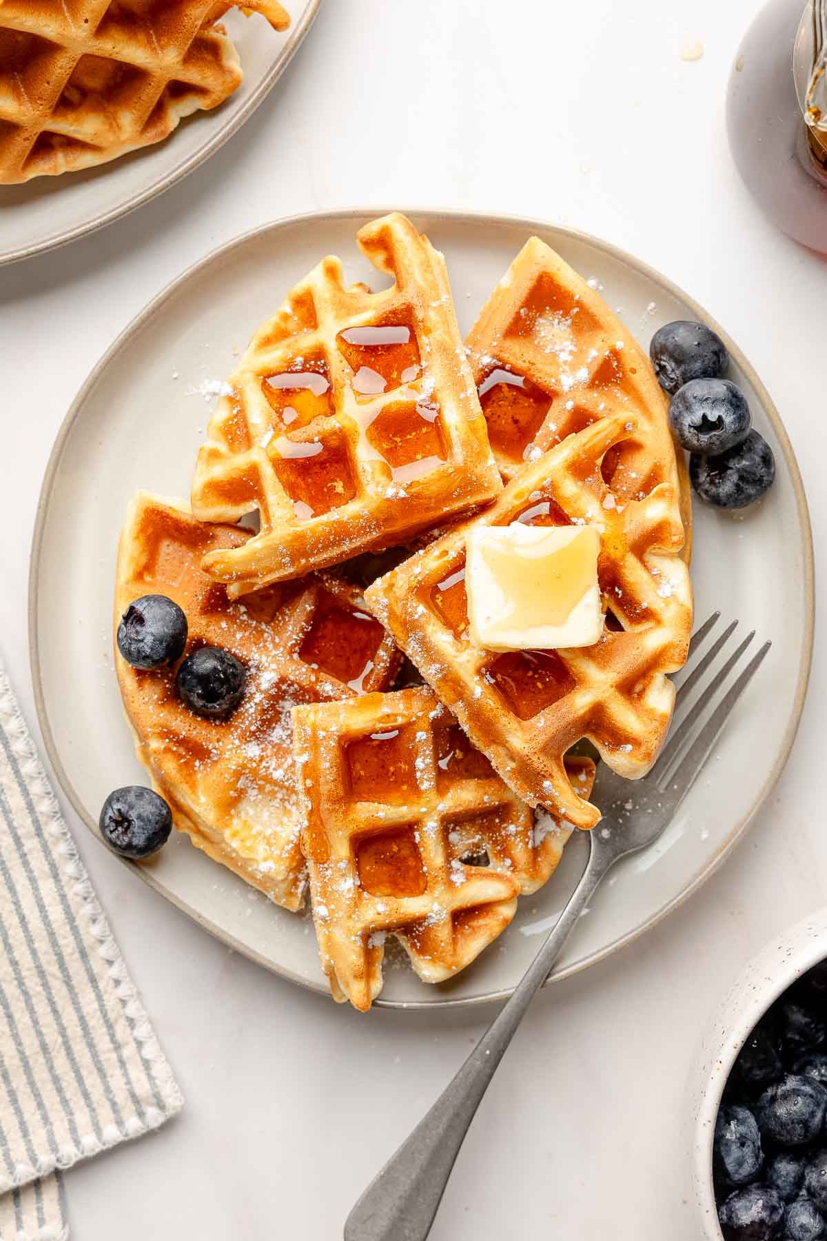 Gluten-free Belgian waffles on a gray plate topped with butter, maple syrup, and powdered sugar, served with fresh blueberries.
