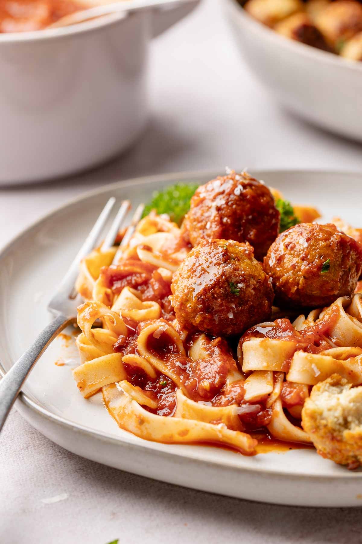 A white plate of gluten-free turkey meatballs served over flat ribbon pasta with rich tomato sauce, garnished with parsley. A fork rests on the left side of the plate, with a Dutch oven and baking dish blurred in the background.