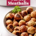 A large shallow white bowl filled with golden-brown baked turkey meatballs, garnished with a sprig of curly parsley. A blurred bowl of pasta and a jar of marinara sauce are visible in the background.