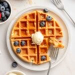 Overhead view of a gluten-free Belgian waffle on a white plate topped with whipped butter, maple syrup, and fresh blueberries, with a bite taken out on a fork.