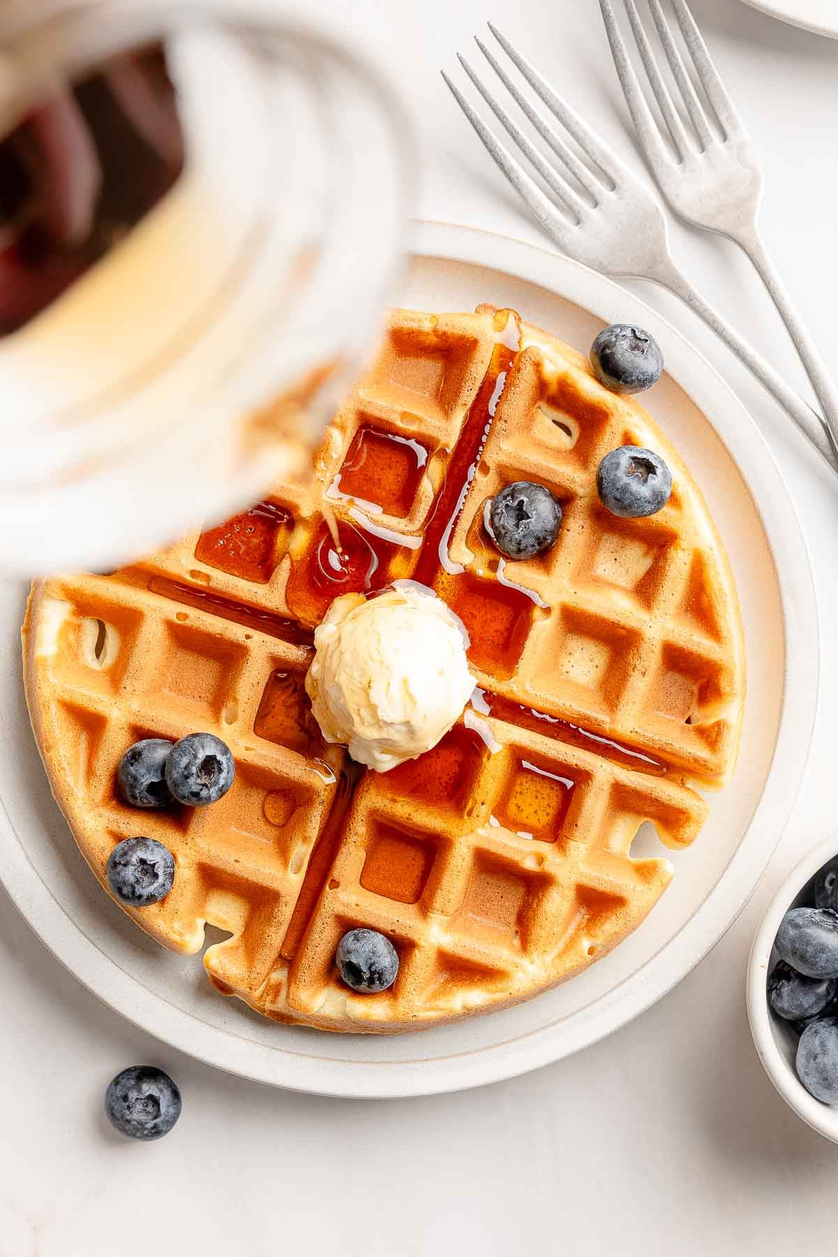 Overhead view of gluten-free waffles garnished with butter and blueberries, is drizzled with syrup.