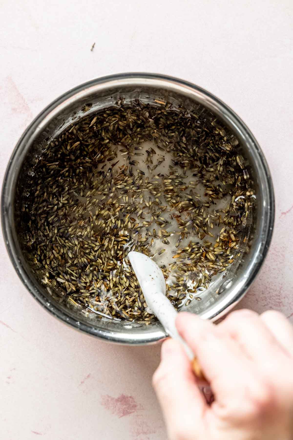 Hand stirring dried lavender buds and dissolved sugar mixture in a saucepan.